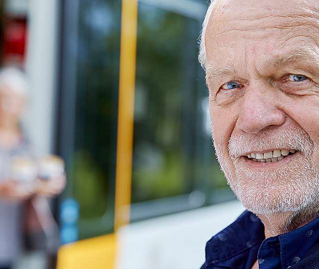 Ein älterer Mann steht an einer Bahnhaltestelle. Verschwommen im Hintergrudn erkennt man eine Frau beim aussteigen aus der Bahn.