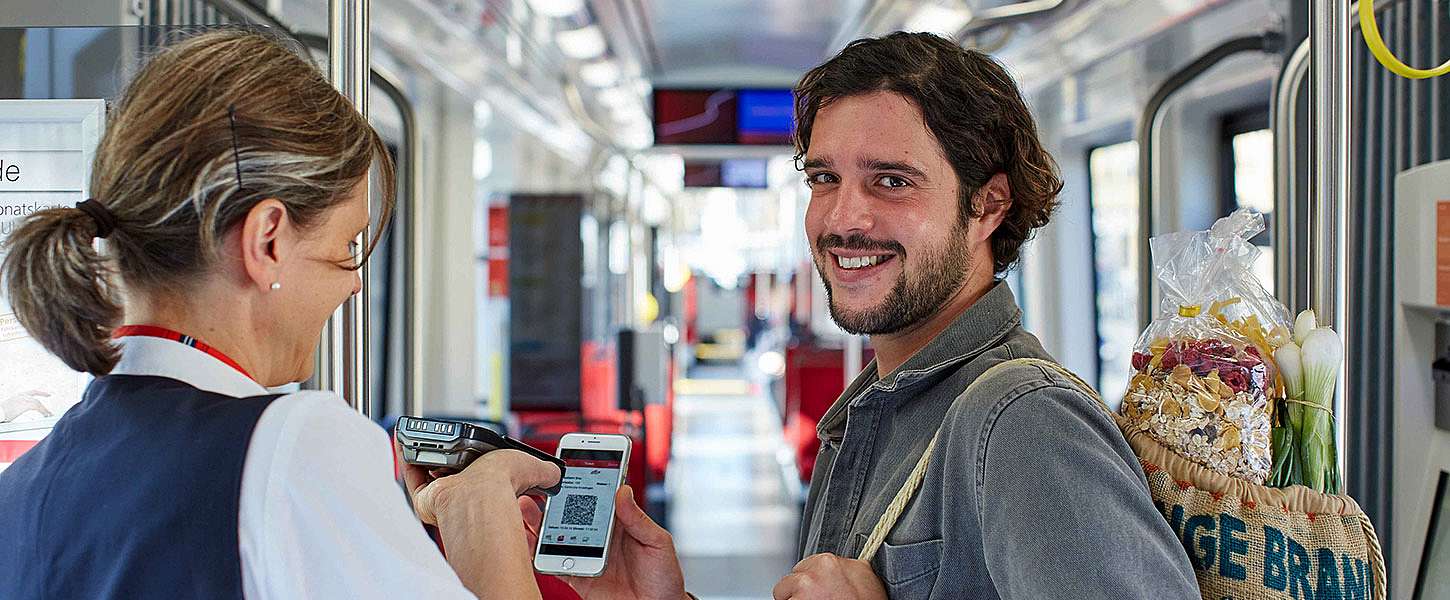 Ein Mann mit einer Tasche voller Lebensmitteln über der Schulter zeigt einer Fahrkartenkontrolleurin seine Fahrkarte auf seinem Smartphone.