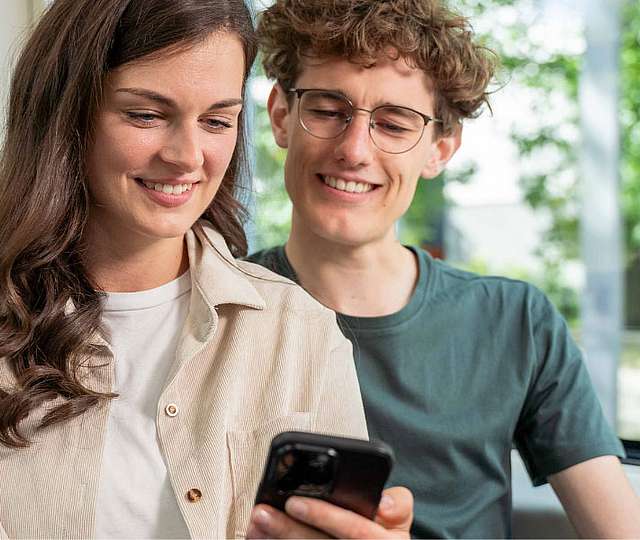 Eine junge Frau und ein junger Mann sitzen in der Bahn und schauen auf ein Smartphone.