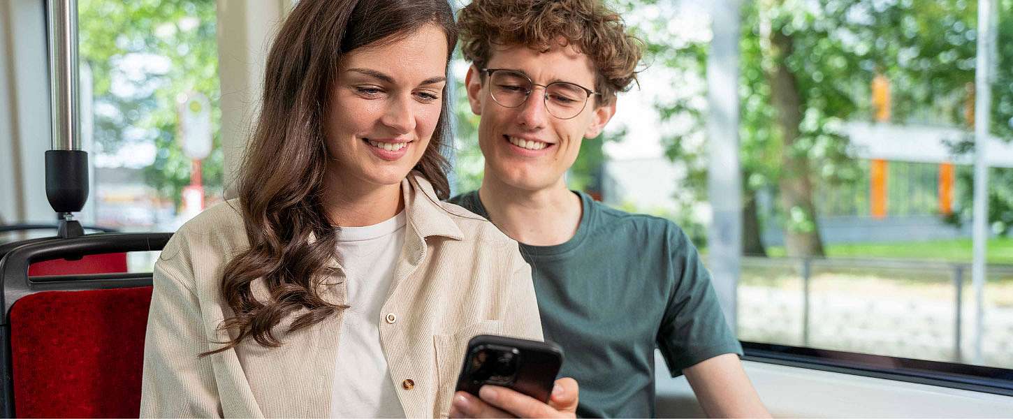 Eine junge Frau und ein junger Mann sitzen in der Bahn und schauen auf ein Smartphone.