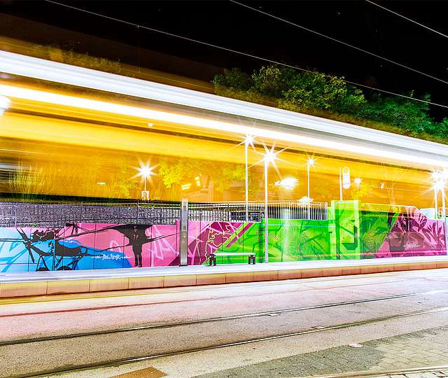 Langzeitbelichtung einer Haltestelle bei Nacht. Die Wände der Haltestelle sind bunt bemalt. Man sieht die Licht-Silhouette einer Bahn durchfahren.