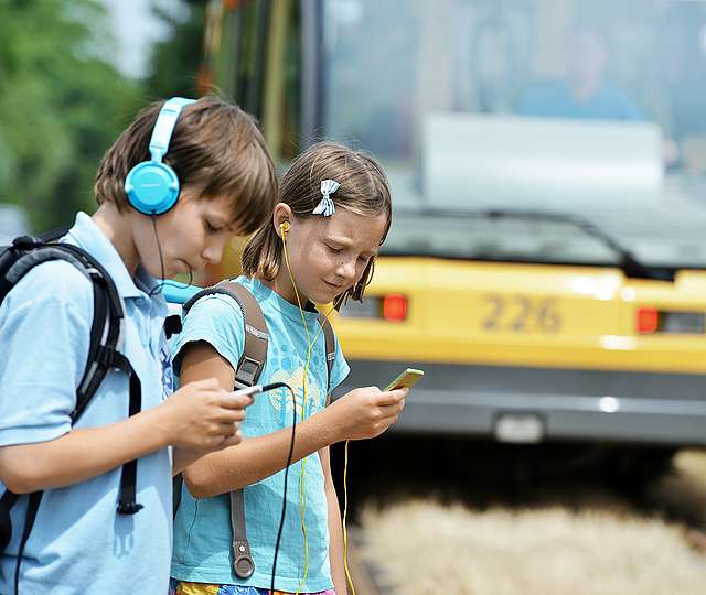 Ein Mädchen und ein Junge mit Schulrucksäcken haben jeweils Kopfhörer auf und schauen auf die Smartphones in ihren Händen. Im Hintergrund ist eine Bahn.