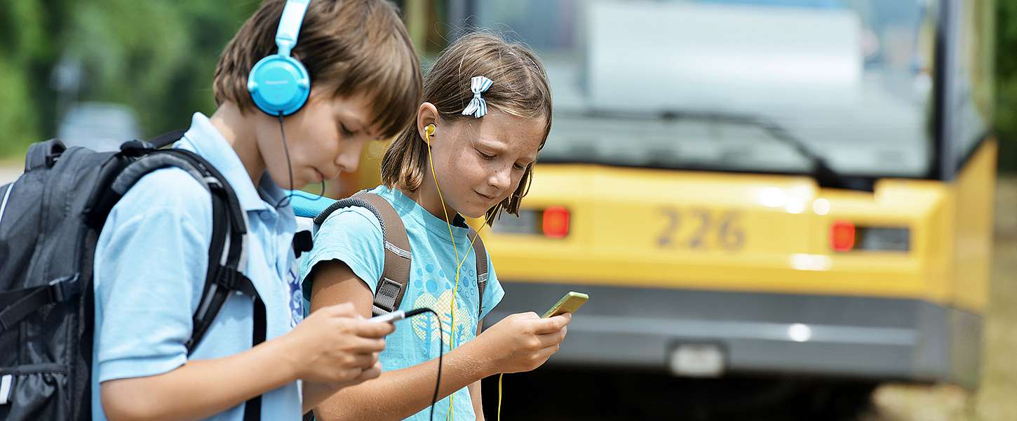 Ein Mädchen und ein Junge mit Schulrucksäcken haben jeweils Kopfhörer auf und schauen auf die Smartphones in ihren Händen. Im Hintergrund ist eine Bahn.