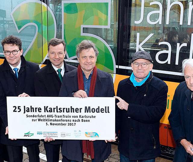 Sechs Männer halten ein Banner. Das Banner bewirbt das Jubiläum 25 Jahre Karlsruher Modell. Hinter den Männern steht eine Bahn, die ebenfalls das Jubiläum bewirbt.