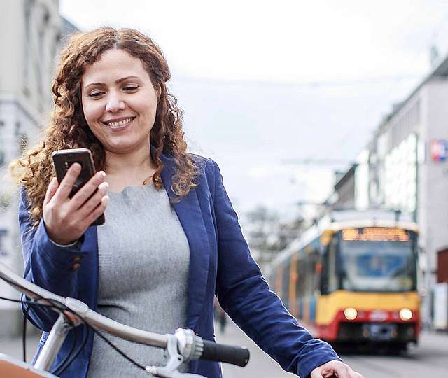 Eine junge Frau schaut lächelnd auf ihr Smartphone. Hinter ihr sieht man eine Bahn.