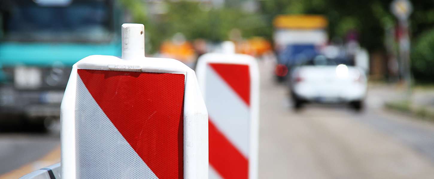 Eine Verkehrsabsperrung an einer Straße. Verschwommen im Hintergrund sieht man ein Auto und einen LKW