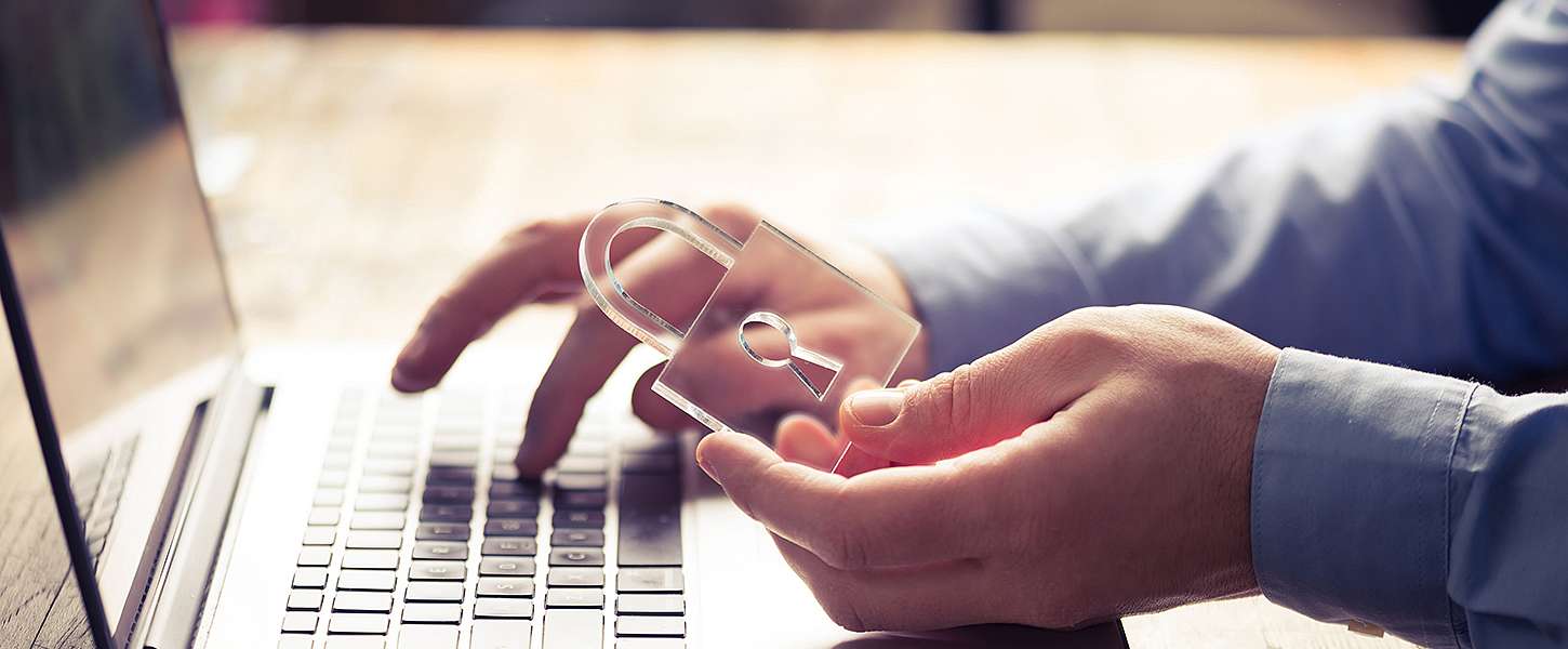 Eine Hand tippt auf dem Laptop, eine zweite Hand hält ein Schloss aus Plastik.