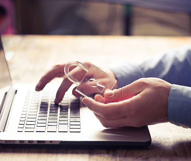 Eine Hand tippt auf dem Laptop, eine zweite Hand hält ein Schloss aus Plastik.