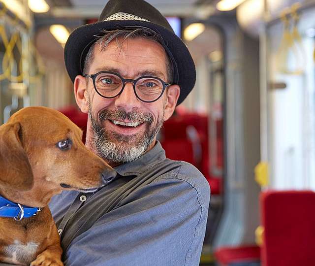 Ein älterer Mann mit Hut und Bart in einer Bahn hält einen Hund auf dem Arm.