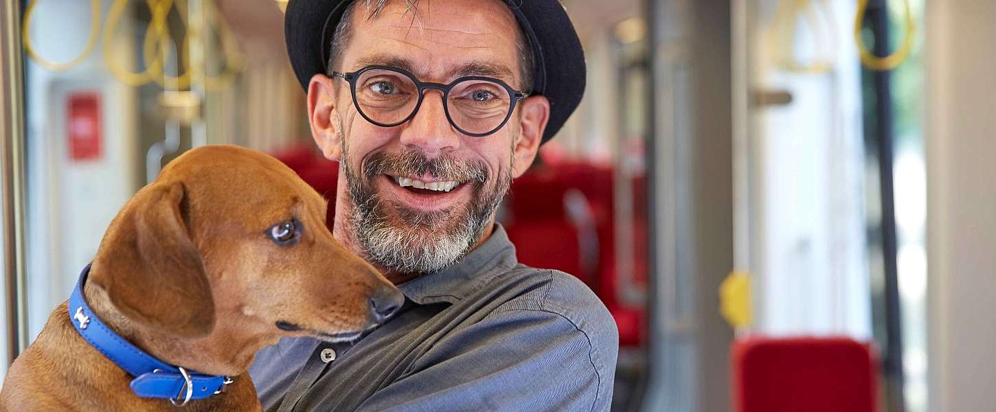 Ein älterer Mann mit Hut und Bart in einer Bahn hält einen Hund auf dem Arm.