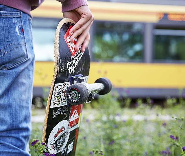 Ein junger Mann, mit der Rückseite zum Betrachter gewandt, hält ein Skateboard in der rechten Hand und schaut vom Gleissteg aus auf die vor ihm stehende gelbe KVV-Bahn.