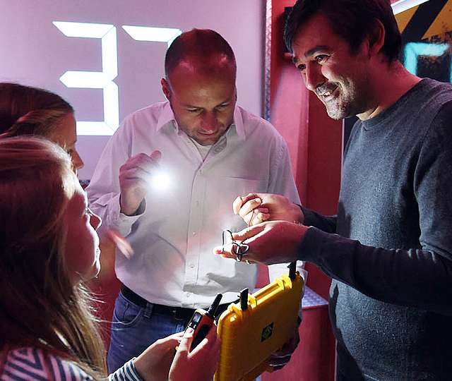 Zwei Männer und zwei Mädchen begutachten Elektronik in einem kleinen gelben Koffer. Im Hintergrund läuft ein Countdown.