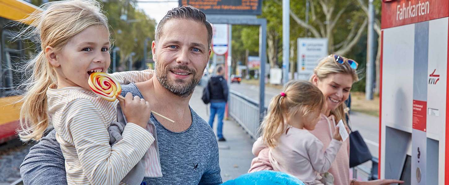 Eine Familie steht an einer Bahnhaltestelle. Der Mann hält ein Mädchen mit Lolli auf dem Arm. Die Frau steht vor dem Fahrkartenautomat. Sie hält ebenfalls ein Mädchen auf dem Arm. Das Mädchen hat einen Fahrschein in der Hand.