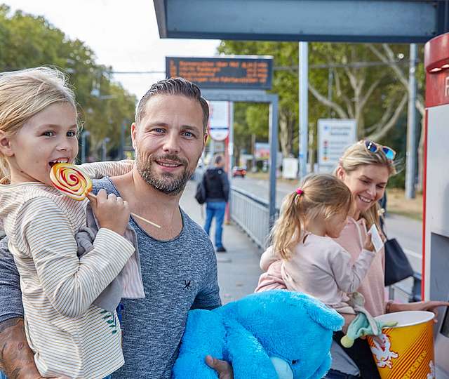 Eine Familie steht an einer Bahnhaltestelle. Der Mann hält ein Mädchen mit Lolli auf dem Arm. Die Frau steht vor dem Fahrkartenautomat. Sie hält ebenfalls ein Mädchen auf dem Arm. Das Mädchen hat einen Fahrschein in der Hand.