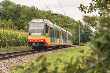 Eine AVG-Stadtbahn fährt auf einem Streckenabschnitt der Kraichtalbahn. Im Hintergrund sind bewaldete Hügel zu sehen.