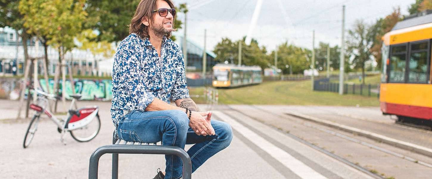 Ein Mann mittleren Alters mit Sonnenbrille sitzt an einem schönen Tag auf einer Bank an einer Bahn-Haltestelle.