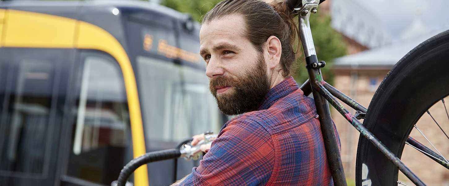 Ein junger Mann mit Bart hat ein Fahrrad über der Schulter. Er steht vor einer Bahn.