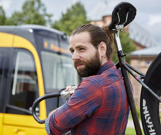 Ein junger Mann mit Bart hat ein Fahrrad über der Schulter. Er steht vor einer Bahn.