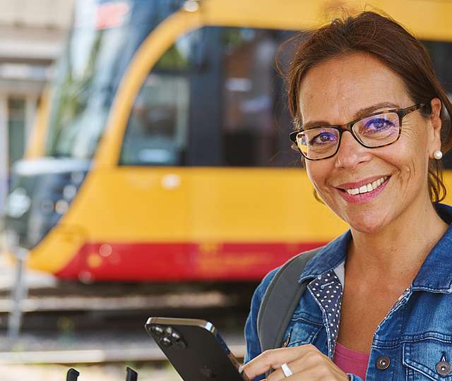 Eine Frau steht vor einem Voi-Roller und hält ihr Smartphone in der Hand. Sie lächelt dabei. Im Hintergrund ist eine Bahn zu sehen.