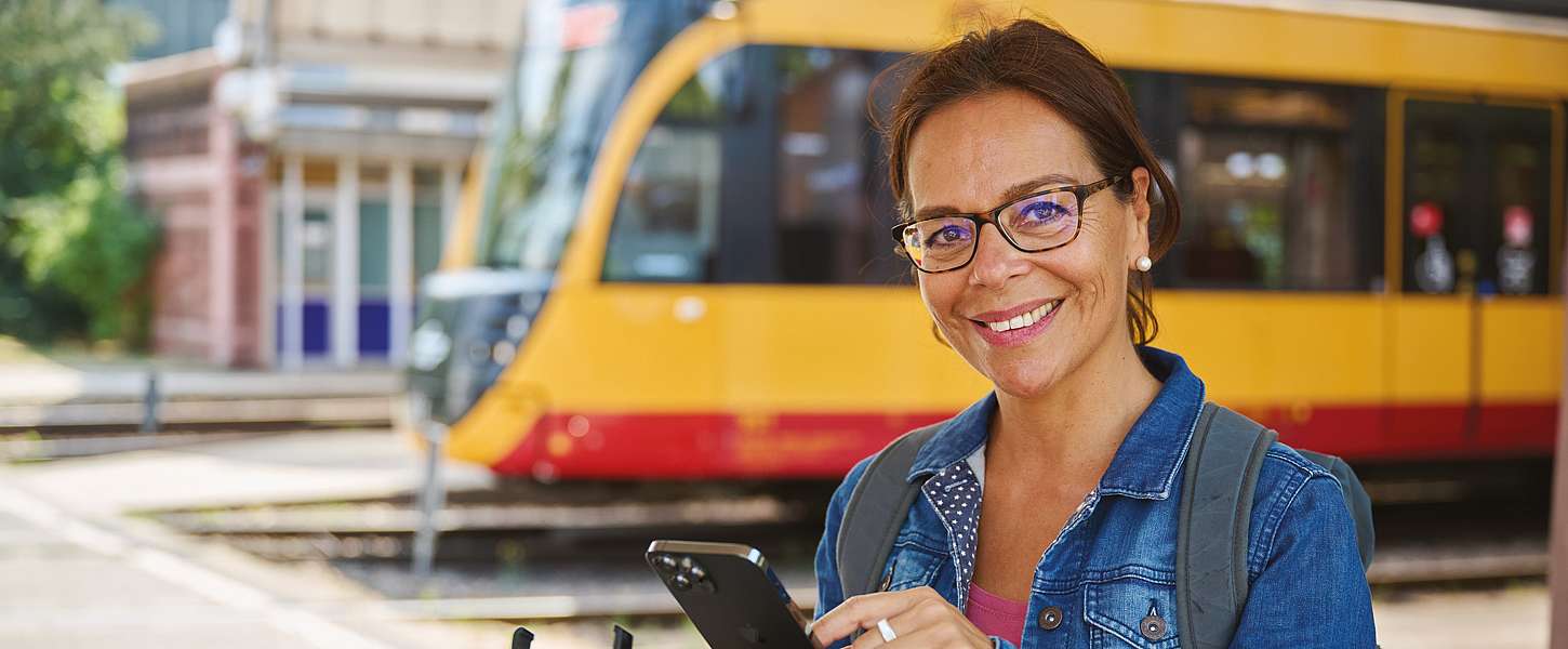 Eine Frau steht vor einem Voi-Roller und hält ihr Smartphone in der Hand. Sie lächelt dabei. Im Hintergrund ist eine Bahn zu sehen.