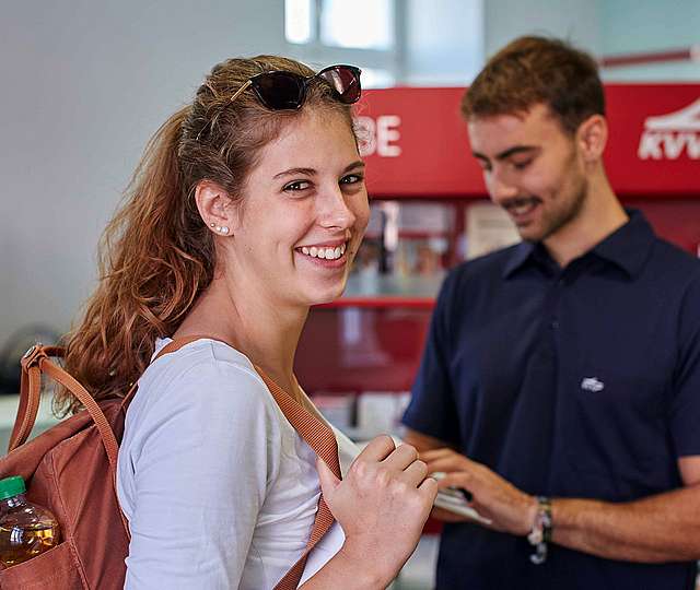 Eine junge Frau mit Rucksack blickt lächelnd in die Kamera. Im Hintergrund sind ein KVV-Mitarbeiter und ein KVV-Prospektständer zu sehen.