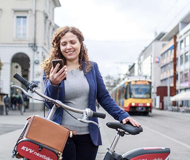 Frau mit Fahrrad schauta auf ihr Smartphone. Im Hintergrund sind Bahnen zu sehen.