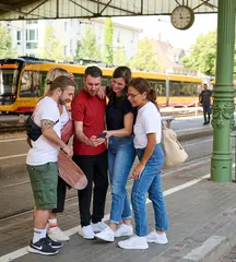 Fünf Menschen stehen in einem Halbkris und blicken gemeinsam auf ein Smartphone. Im Hintergrund ist eine Stadtbahn zu sehen.
