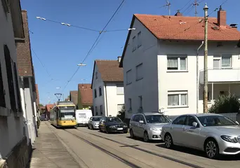 Eine Trambahn der VBK fährt durch den Ortskern von Daxlanden. Neben dem Bahngleis stehende parkende Autos. Zudem sind Häuser zu sehen.
