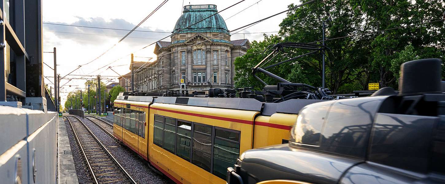 Eine ältere Frau und ein älterer Mann stehen in einer Bahn neben einem Fahrkartenentwerter. Die Frau lächelt. Sie hat einen Arm auf dem Entwerter abgelegt. Neben ihrer Hand steht ein To Go Becher.