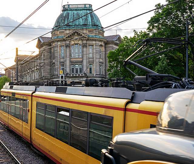 Eine ältere Frau und ein älterer Mann stehen in einer Bahn neben einem Fahrkartenentwerter. Die Frau lächelt. Sie hat einen Arm auf dem Entwerter abgelegt. Neben ihrer Hand steht ein To Go Becher.