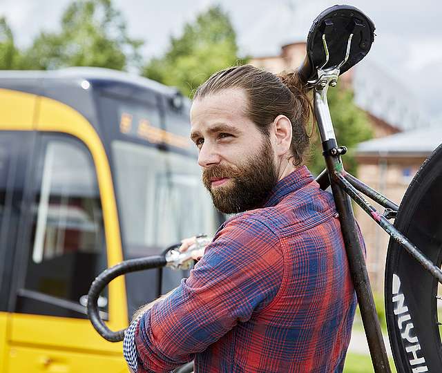 Ein junger Mann in karriertem Hemd trägt ein Fahrrad über der Schulter. Im Hintergrund sieht man eine Bahn der KVV.