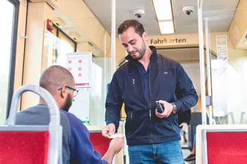 Ein Fahrausweisprüfer hält ein Prüfgerät in der Hand und schaut sich in einer Bahn den Fahrschein eines Fahrgastes an.