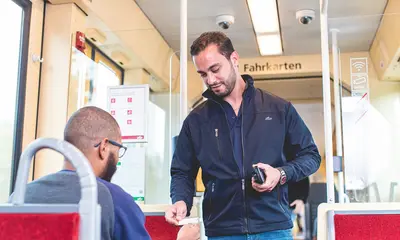 Ein Fahrausweisprüfer hält ein Prüfgerät in der Hand und schaut sich in einer Bahn den Fahrschein eines Fahrgastes an.