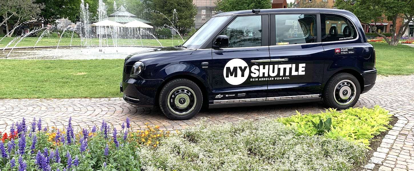 Ein  London Taxi mit der MyShuttle Beklebung steht an einem schönen Platz mit Springbrunnen.